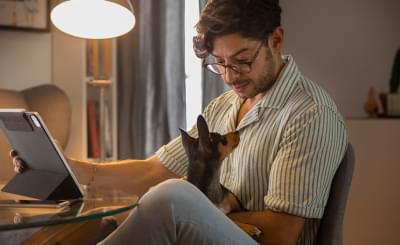 Imagem mostra um homem de óculos sentado em casa trabalhando em um tablet com seu cachorro no colo