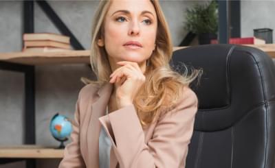 Image shows a white woman with light, straight hair, wearing formal clothes, sitting in an office, looking to the right with her hand under her chin